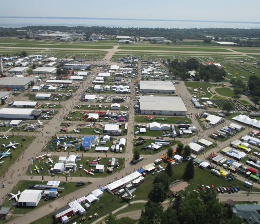 Low-level aerial view of AirVenture, looking down the main thoroughfare toward Boeing Plaza, with the flightline and runway 18/36 beyond. [Credit: LeRoy Cook]